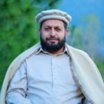 A man outdoors wearing a Chitrali cap, wrapped in a shawl and posing with a serene smile.